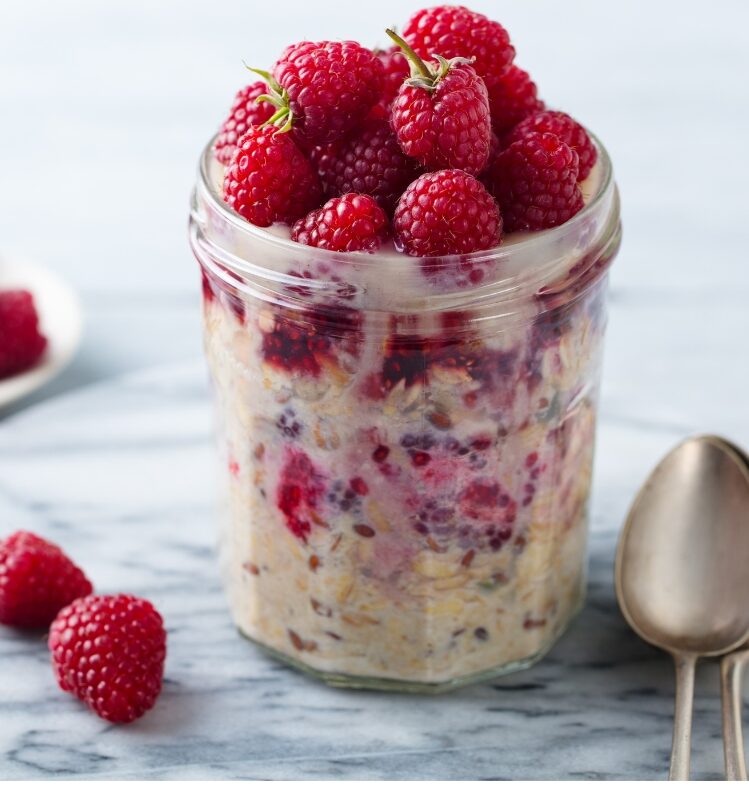 Glass filled bircher muesli, topped with apple and raspberries with a spoon laying next to it