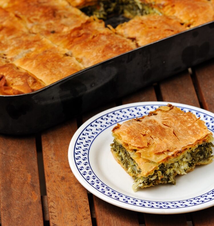 A slice of spanakopita on a plate next to a large dish full of spanakopita